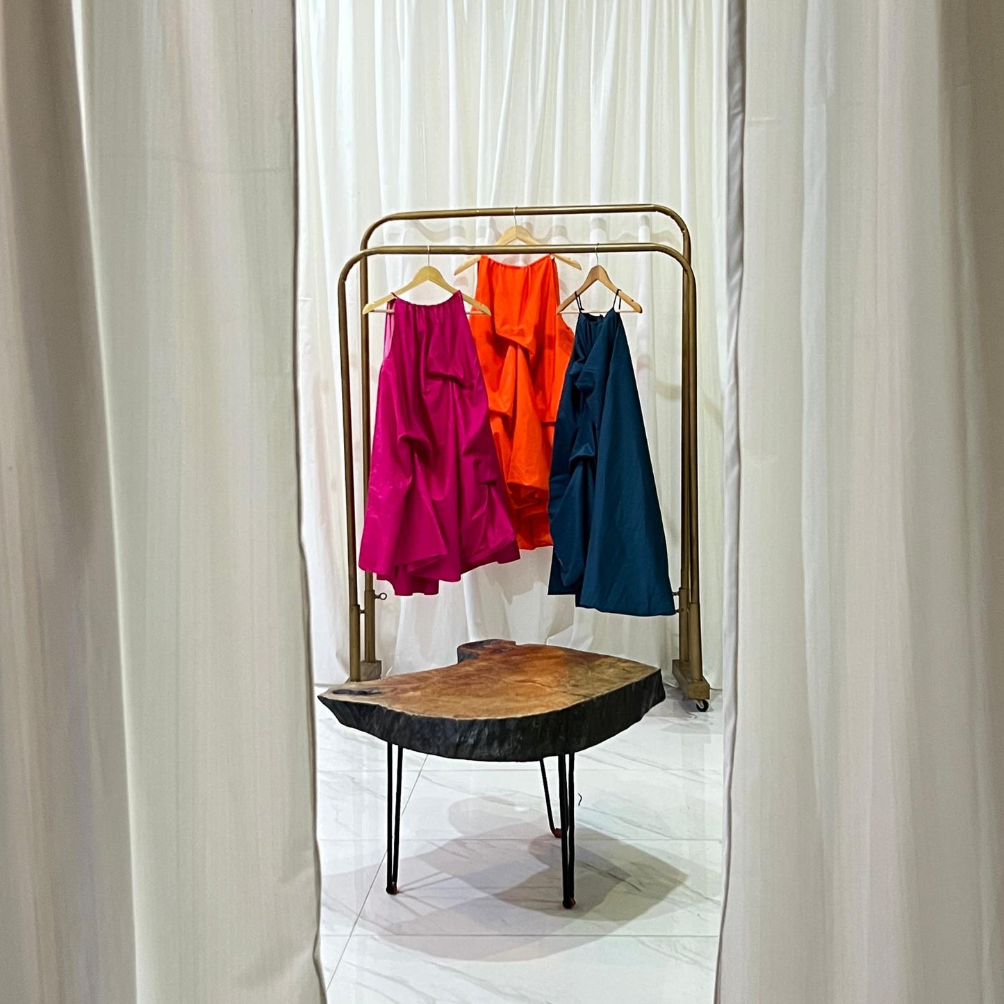 Three colorful dresses hanging on a rack with white curtains on either side.